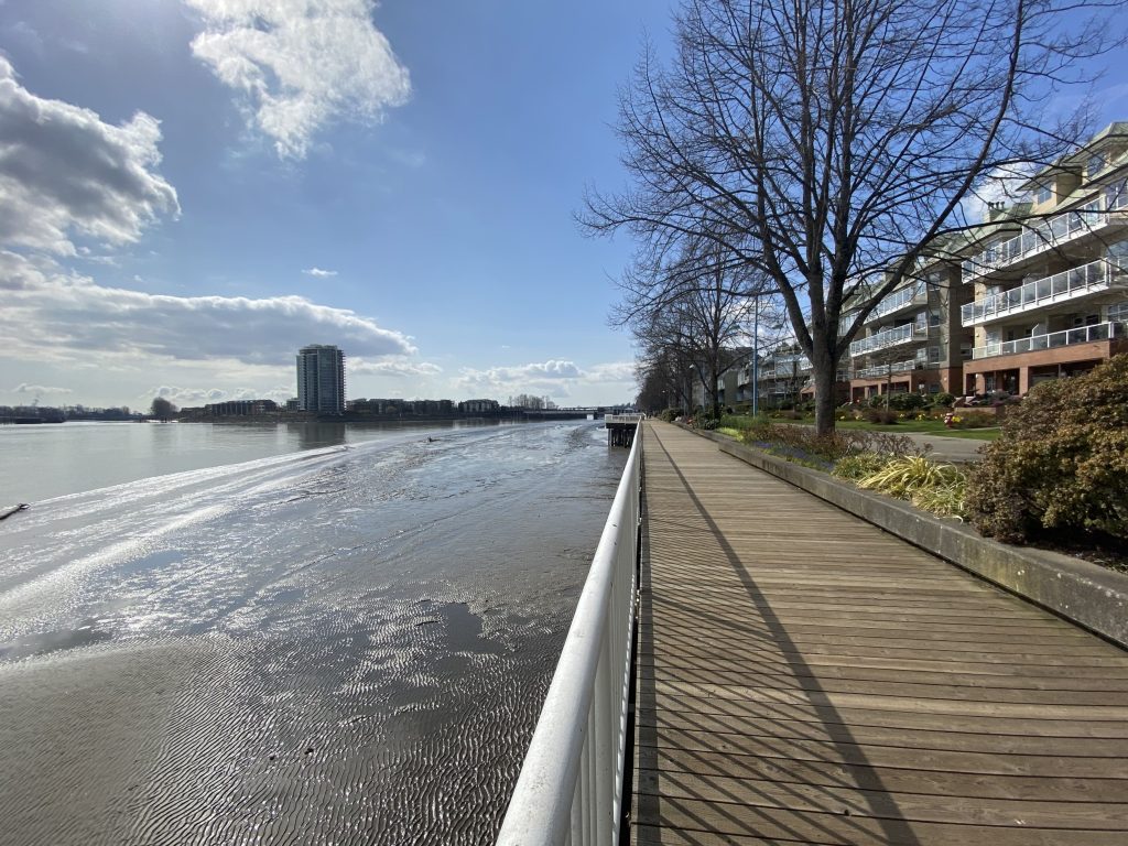 Sunny boardwalk scenery with the Fraser River to the left, the blue sky with some clouds and the boardwalk and apartments to the right.